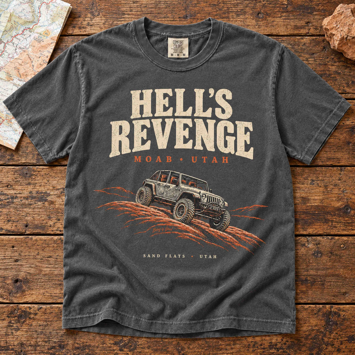 Pepper Hell’s Revenge graphic t-shirt laid flat on a vintage wooden table, featuring an off-road vehicle on orange slickrock.