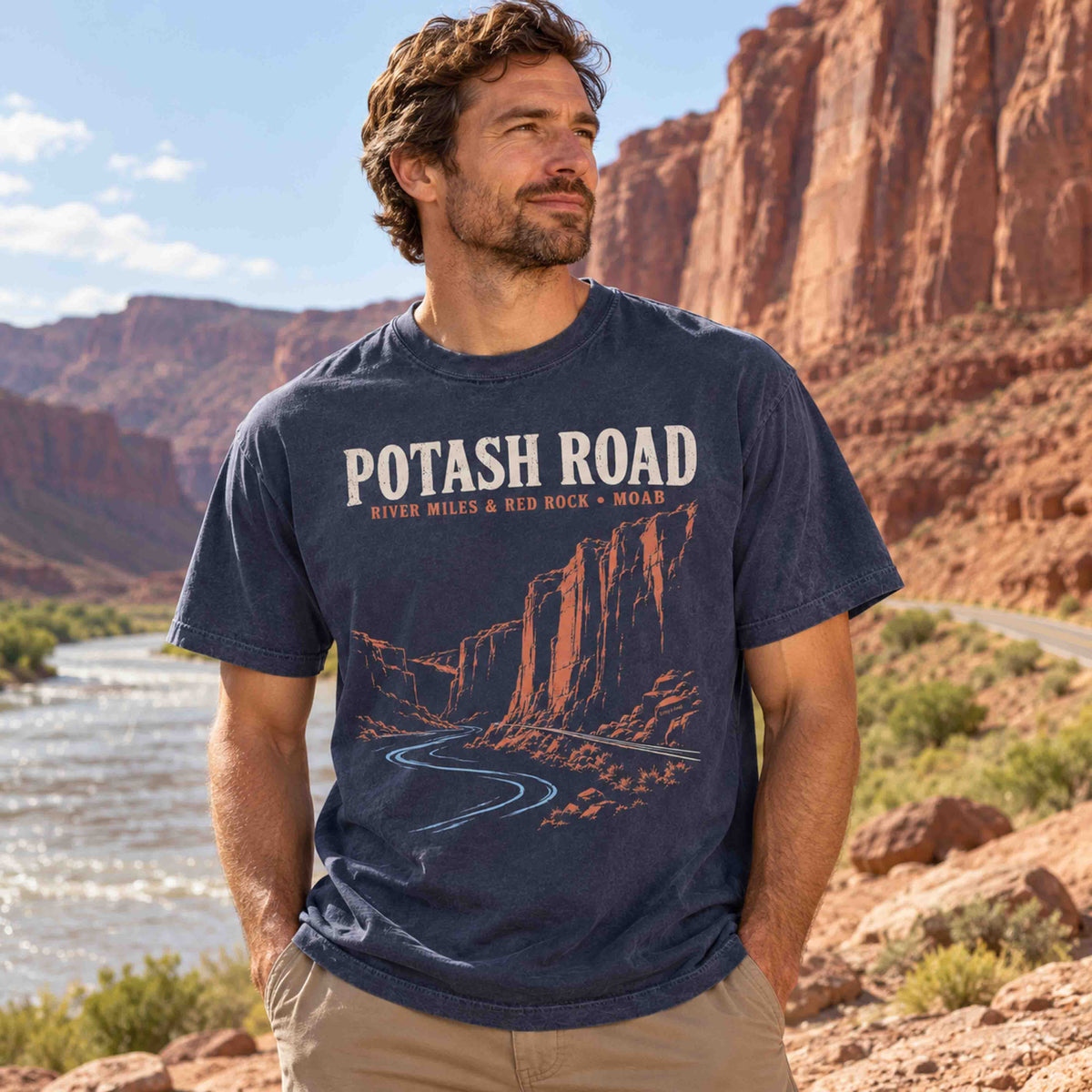 Man wearing a Navy Being is Good Potash Road graphic T-shirt beside a river and red rock canyon walls.