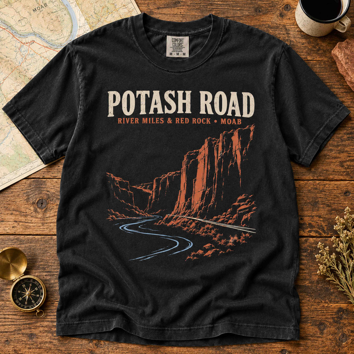 Black Potash Road Moab graphic T-shirt laid flat on rustic wood beside a map, compass and coffee cup.