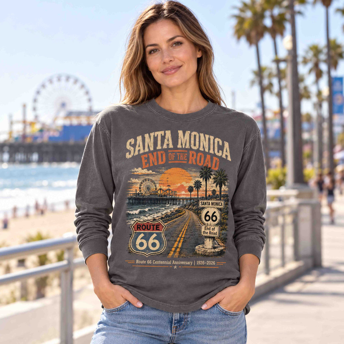 Woman wearing a Pepper Santa Monica Route 66 long sleeve graphic tee on a coastal walkway beside palm trees and the pier.