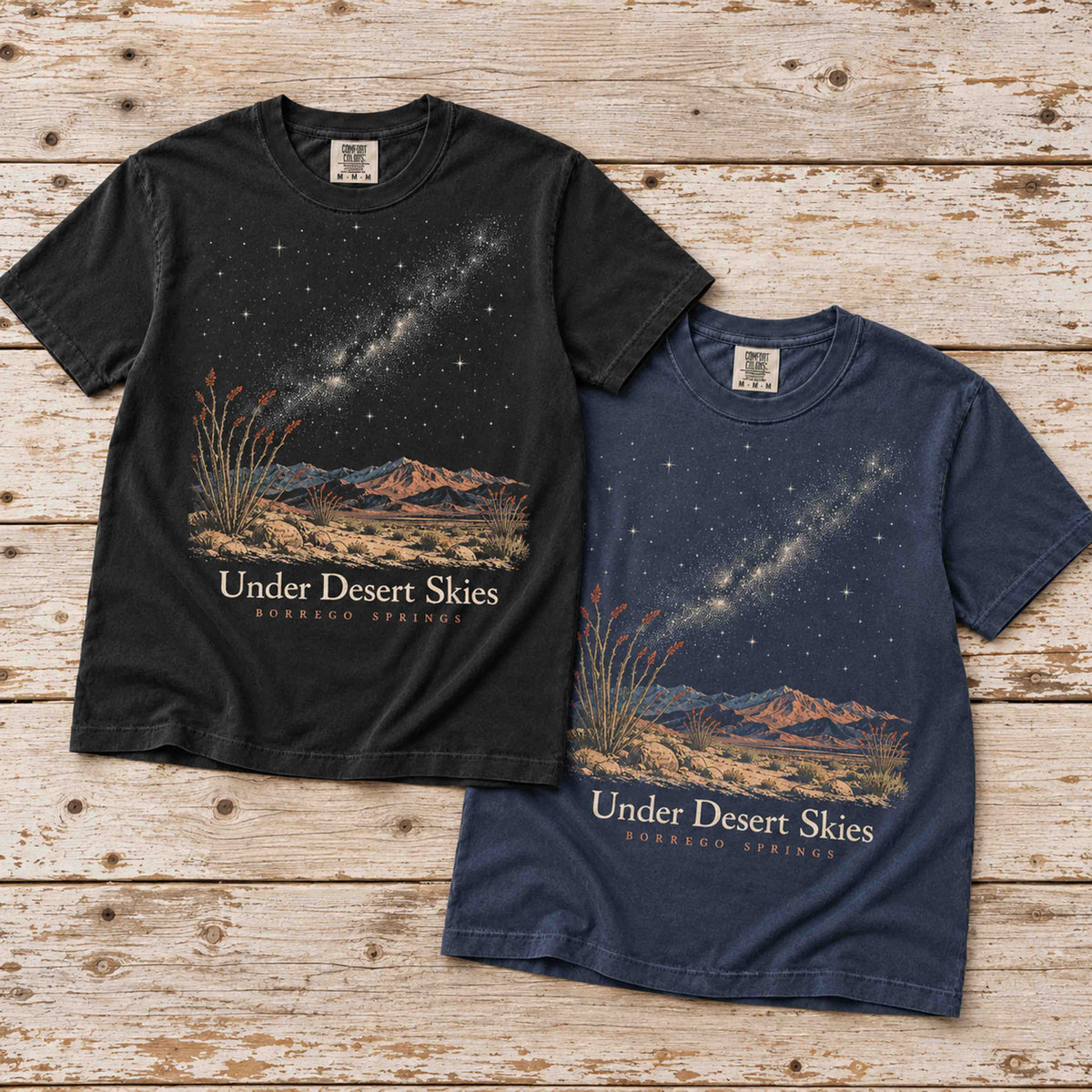 Black and Navy Under Desert Skies Borrego Springs T-shirts overlapping on a weathered wooden surface.