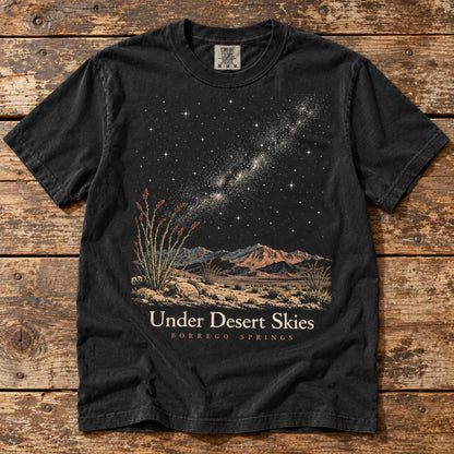 Black Under Desert Skies Borrego Springs T-shirt laid flat on wood, showing Milky Way, mountain and ocotillo artwork.