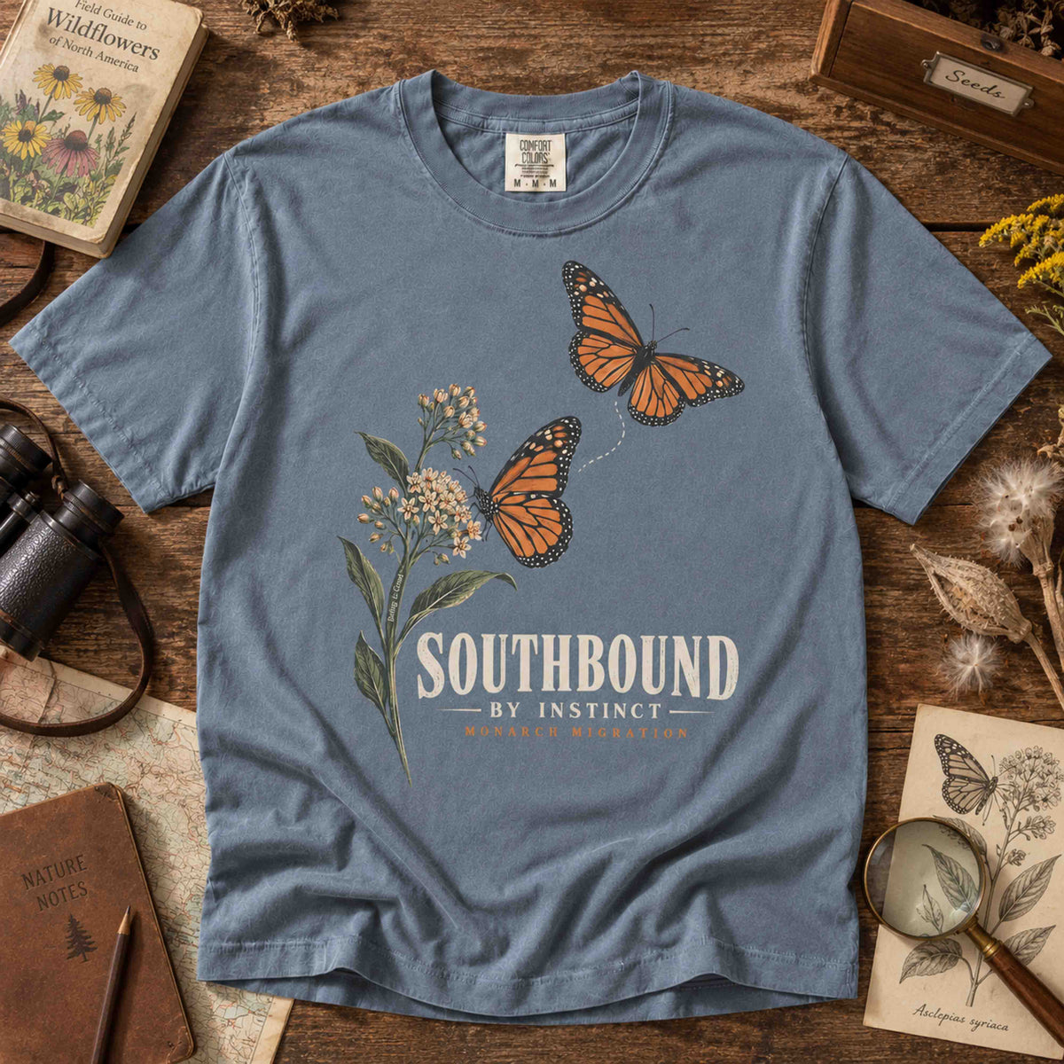 Blue Jean Southbound by Instinct T-shirt laid flat on wood, showing monarch butterflies and flowering botanical artwork.