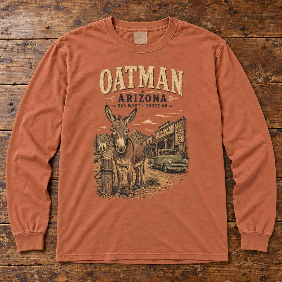 Yam Oatman Arizona Route 66 long sleeve tee laid flat on wood, showing burro, vintage truck and storefront artwork.