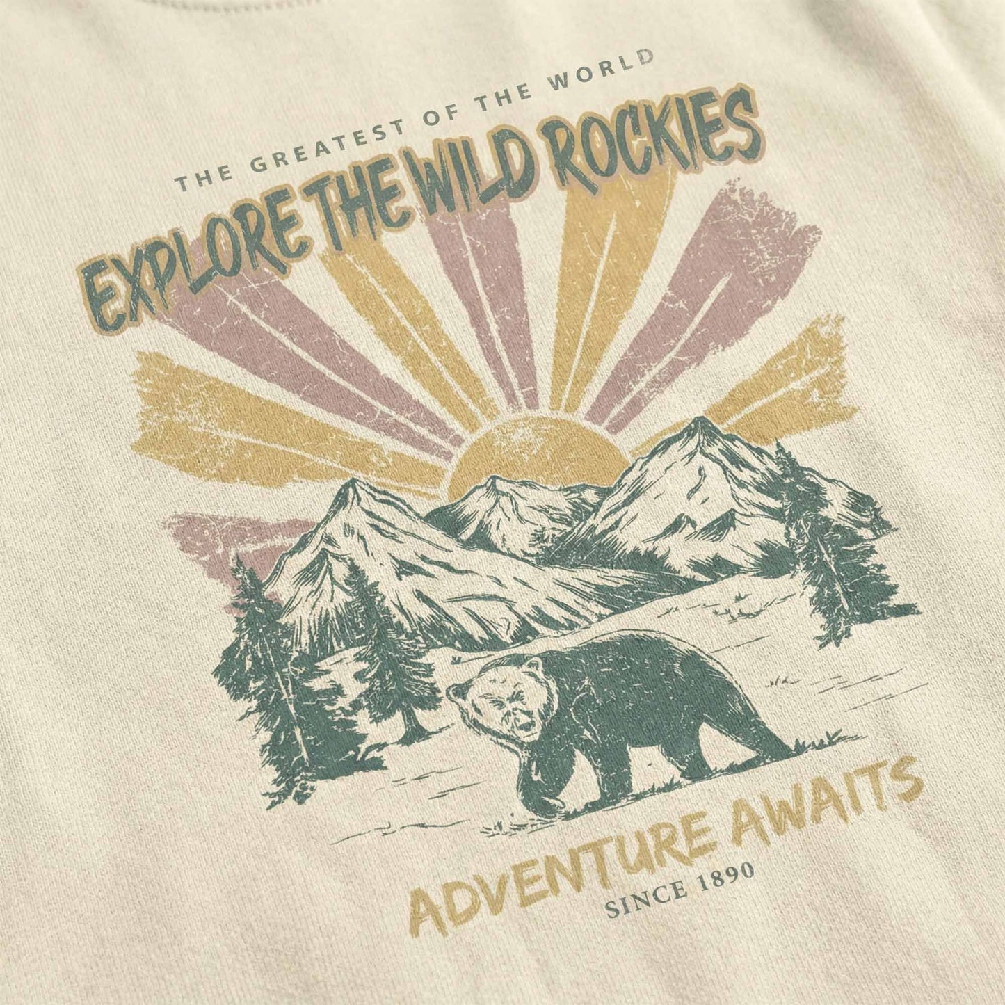 Close-up of a vintage-style graphic on a cream t-shirt featuring a bear, mountains, sun rays, and the text "Explore the Wild Rockies" and "Adventure Awaits".