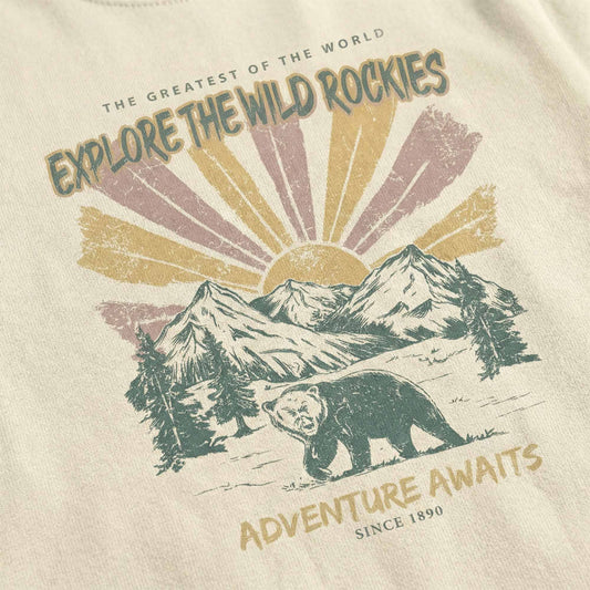 Close-up of a vintage-style graphic on a cream t-shirt featuring a bear, mountains, sun rays, and the text "Explore the Wild Rockies" and "Adventure Awaits".