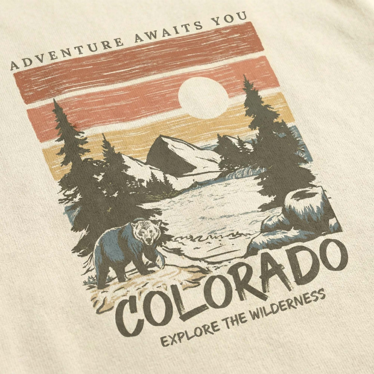 Close-up of a minimal-vintage Colorado graphic on a cream t-shirt, featuring a bear, mountains, pine trees, and the text "Adventure Awaits You" and "Explore the Wilderness".
