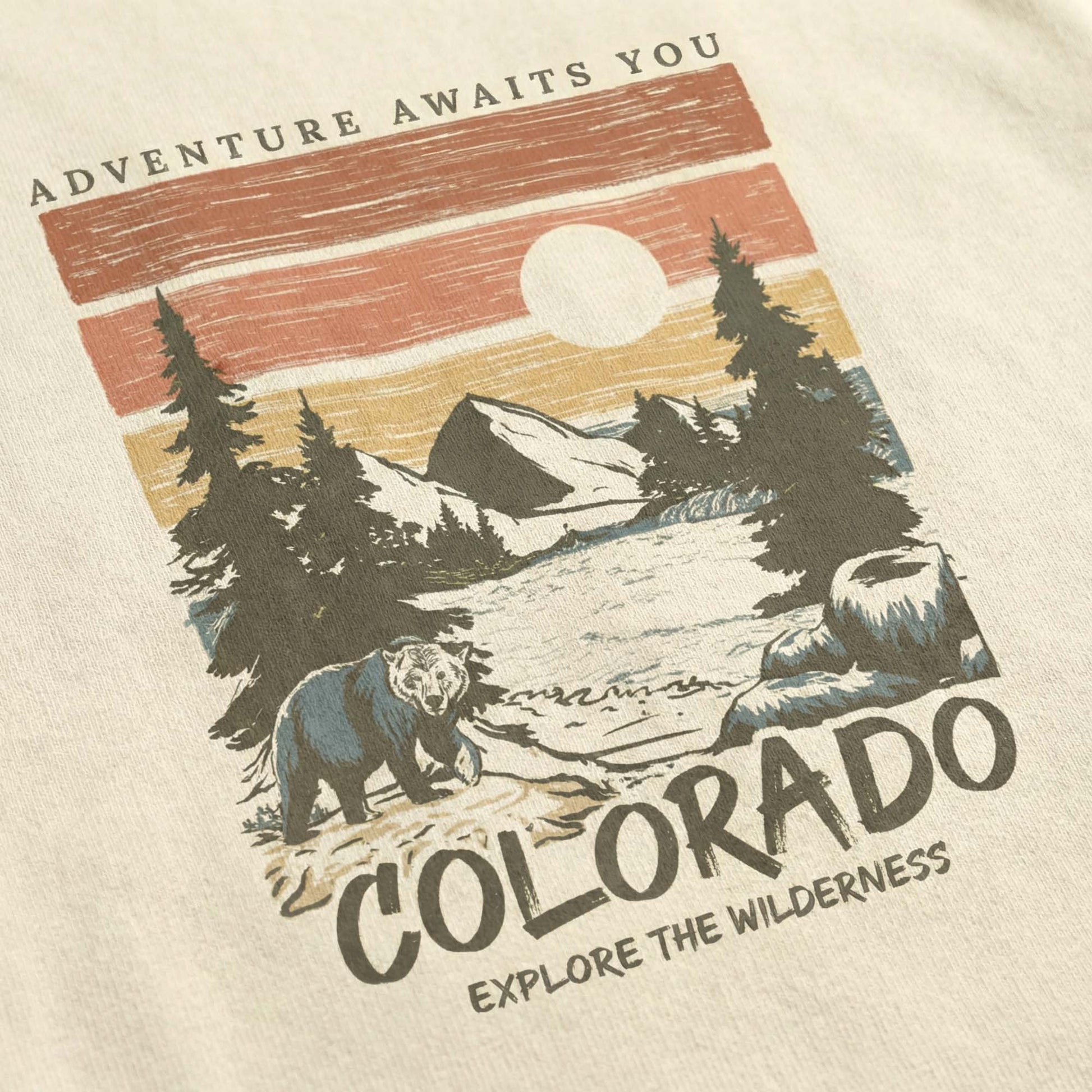 Close-up of a minimal-vintage Colorado graphic on a cream t-shirt, featuring a bear, mountains, pine trees, and the text "Adventure Awaits You" and "Explore the Wilderness".
