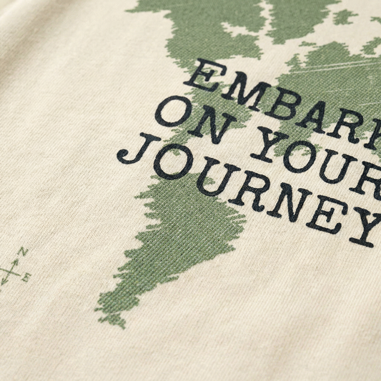 Close-up of a minimal-vintage graphic on cream fabric, featuring a faded green landmass map and the text "Embark On Your Journey".
