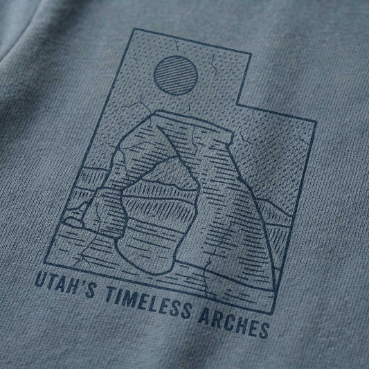 Close-up of a minimal-vintage dark blue graphic on a dusty blue t-shirt, featuring the state outline of Utah, a desert arch, a sun, and the text "Utah's Timeless Arches".