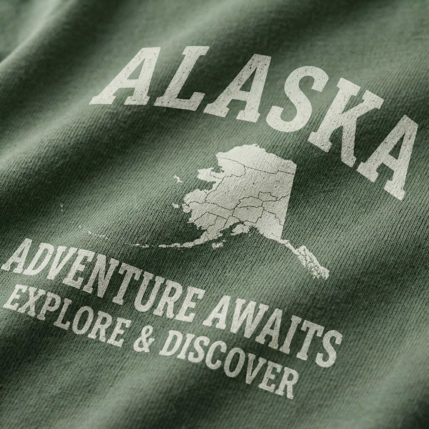 Close-up of a minimal-vintage graphic on green fabric, featuring the state of Alaska and text reading "Alaska", "Adventure Awaits", and "Explore & Discover".