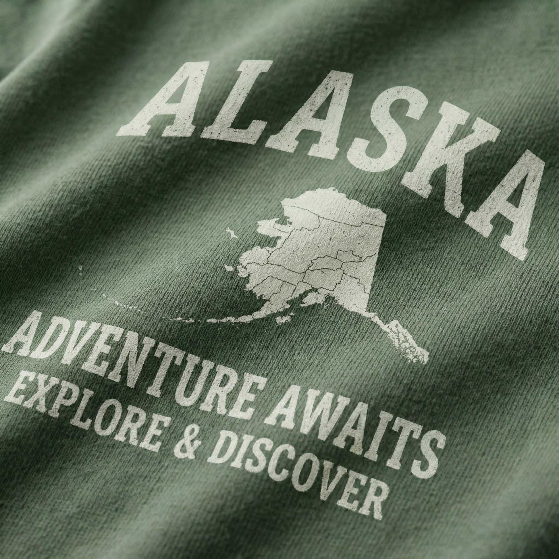 Close-up of a minimal-vintage graphic on green fabric, featuring the state of Alaska and text reading "Alaska", "Adventure Awaits", and "Explore & Discover".