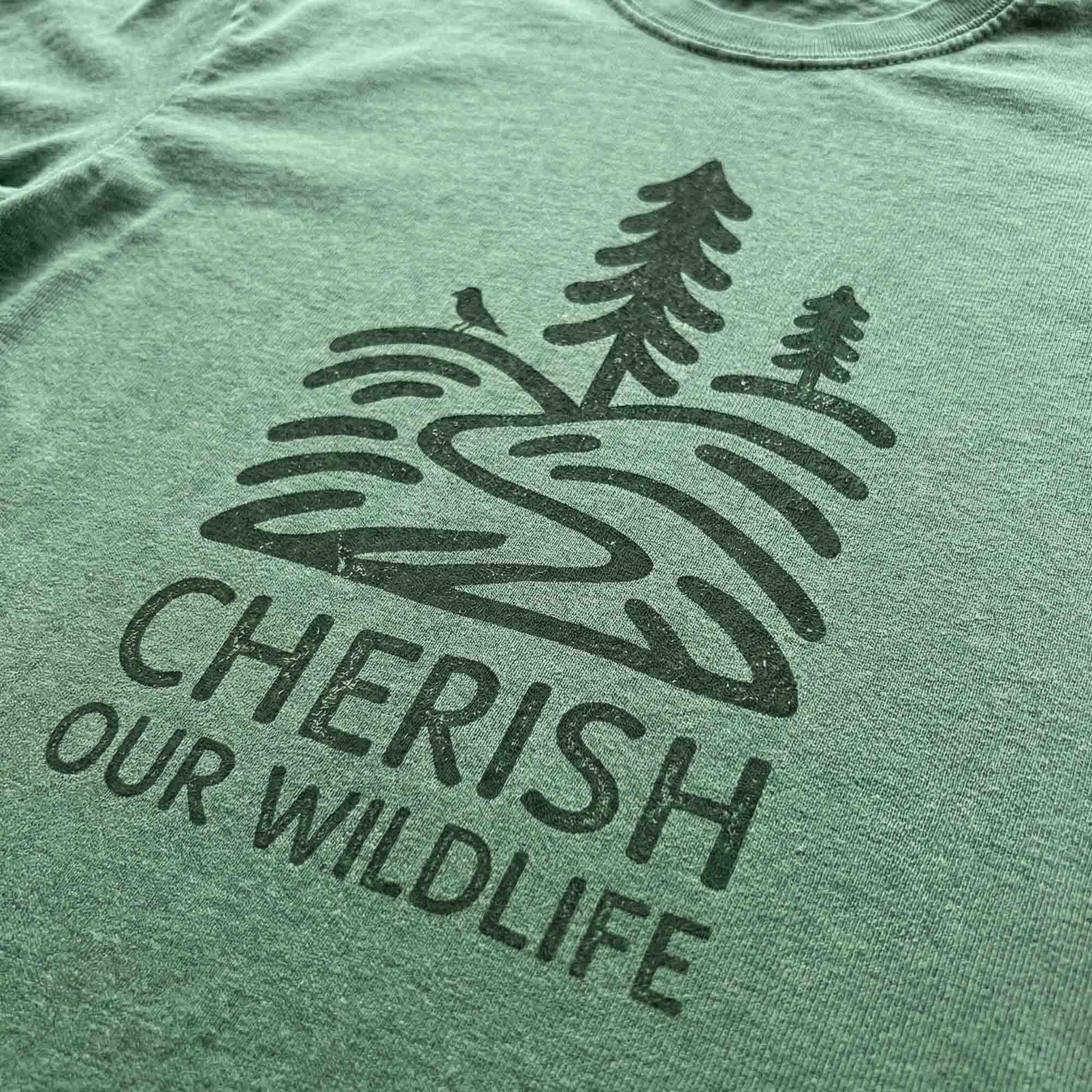 Close-up of a dark green minimal-vintage graphic on a green t-shirt, featuring pine trees, a winding river, and the text "Cherish Our Wildlife".