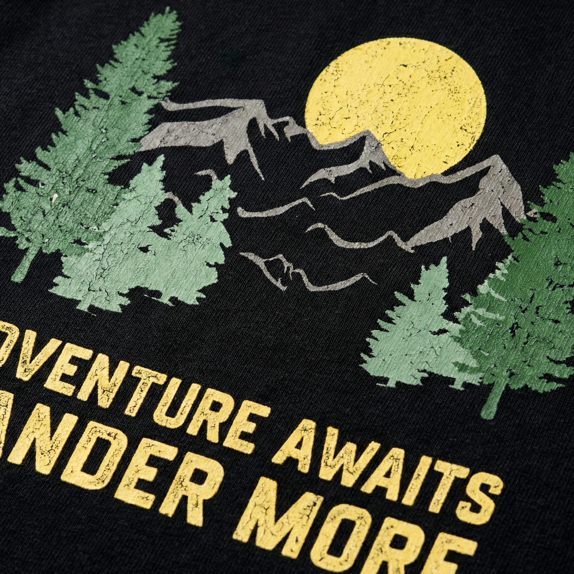 Close-up of a retro outdoor graphic on black fabric, featuring green pine trees, mountains, a yellow sun, and the text "Adventure Awaits Wander More".