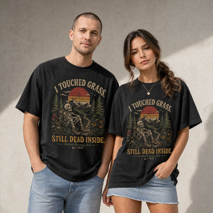 Models wearing black vintage graphic tees featuring a skeleton sitting outdoors with mountains, pine trees, wildflowers, sunset, and text reading I Touched Grass Still Dead Inside.