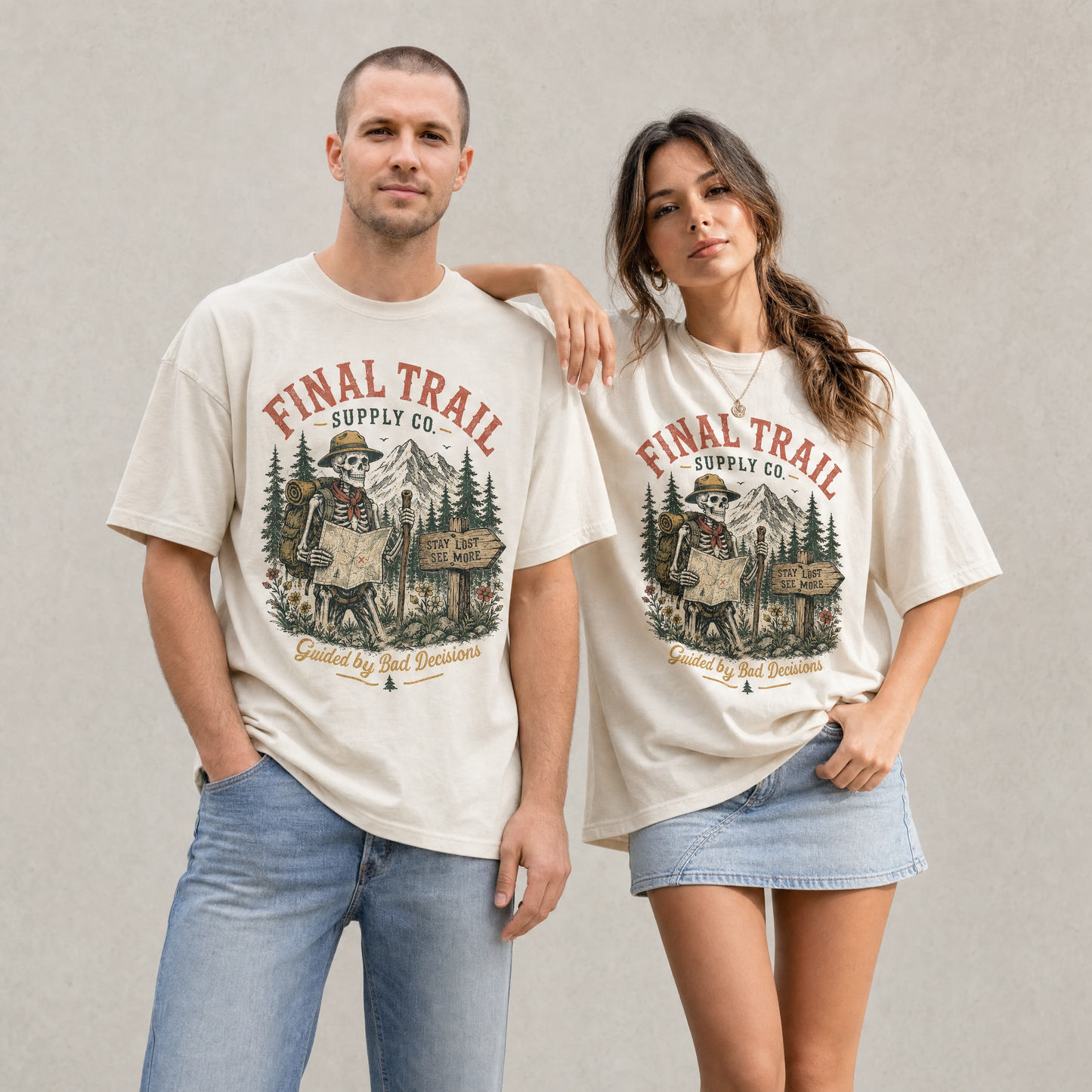 Man and woman wearing sage and ivory graphic t-shirts featuring a skeleton trail guide with mountains, trees, and vintage outdoor artwork