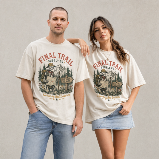 Man and woman wearing sage and ivory graphic t-shirts featuring a skeleton trail guide with mountains, trees, and vintage outdoor artwork