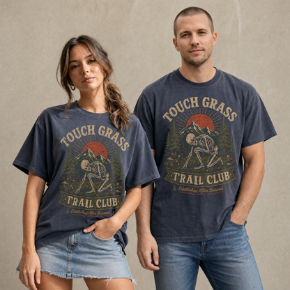 Vintage skeleton outdoor graphic tee featuring a skeleton touching grass with mountains, pine trees, wildflowers, sunset, and text reading Touch Grass Trail Club.