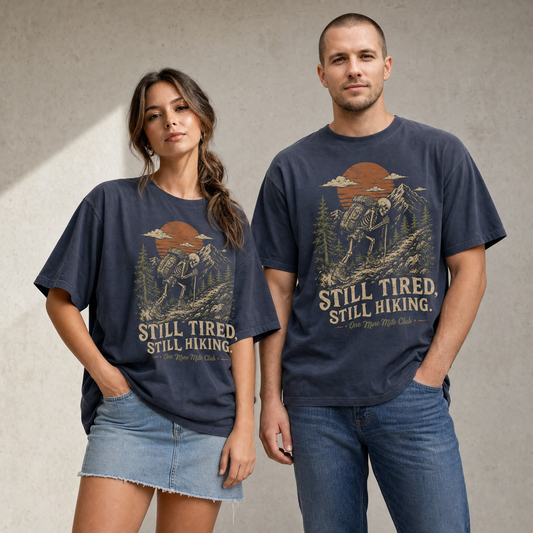 Vintage skeleton hiking graphic tee featuring a tired skeleton backpacking uphill through mountains, pine trees, rocks, sunset, and text reading Still Tired Still Hiking.